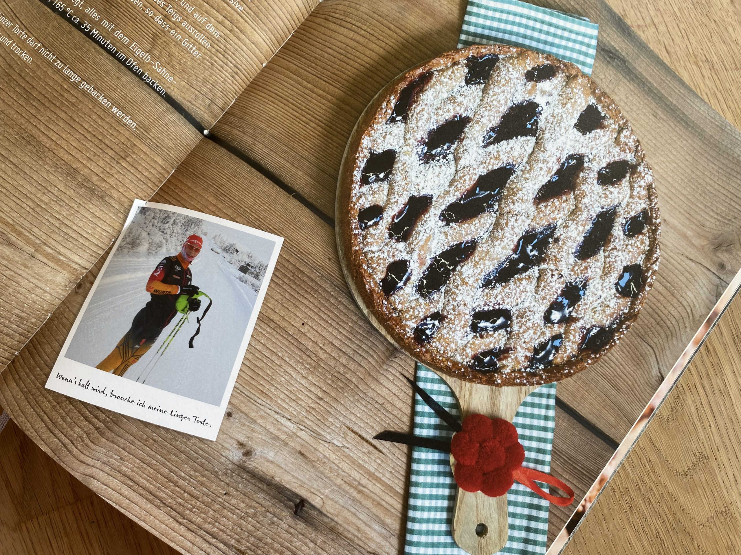 „Mockup eines Kochbuch-Layouts: Doppelseite mit Rezepttext und einer persönlichen Fotokarte auf der linken Seite, gegenüberliegend das Food-Styling einer Linzer Torte auf einem Holzbrett.“