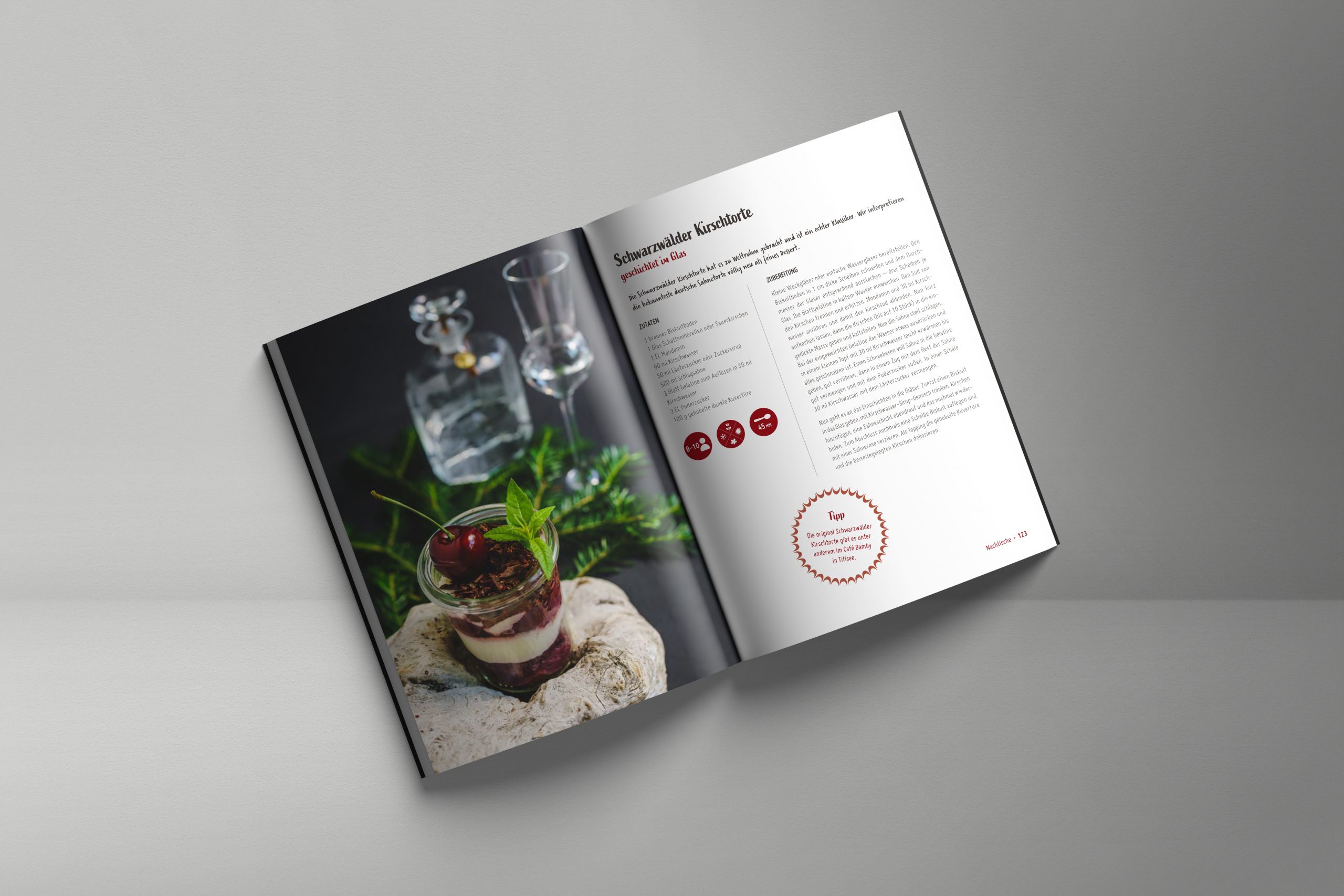 Offenes Rezeptbuch mit Schwarzwälder Kirschtorte im Glas, dekoriert mit frischen Kirschen und Minze, daneben Gläser mit Kirschwasser und Zutatenliste im Hintergrund