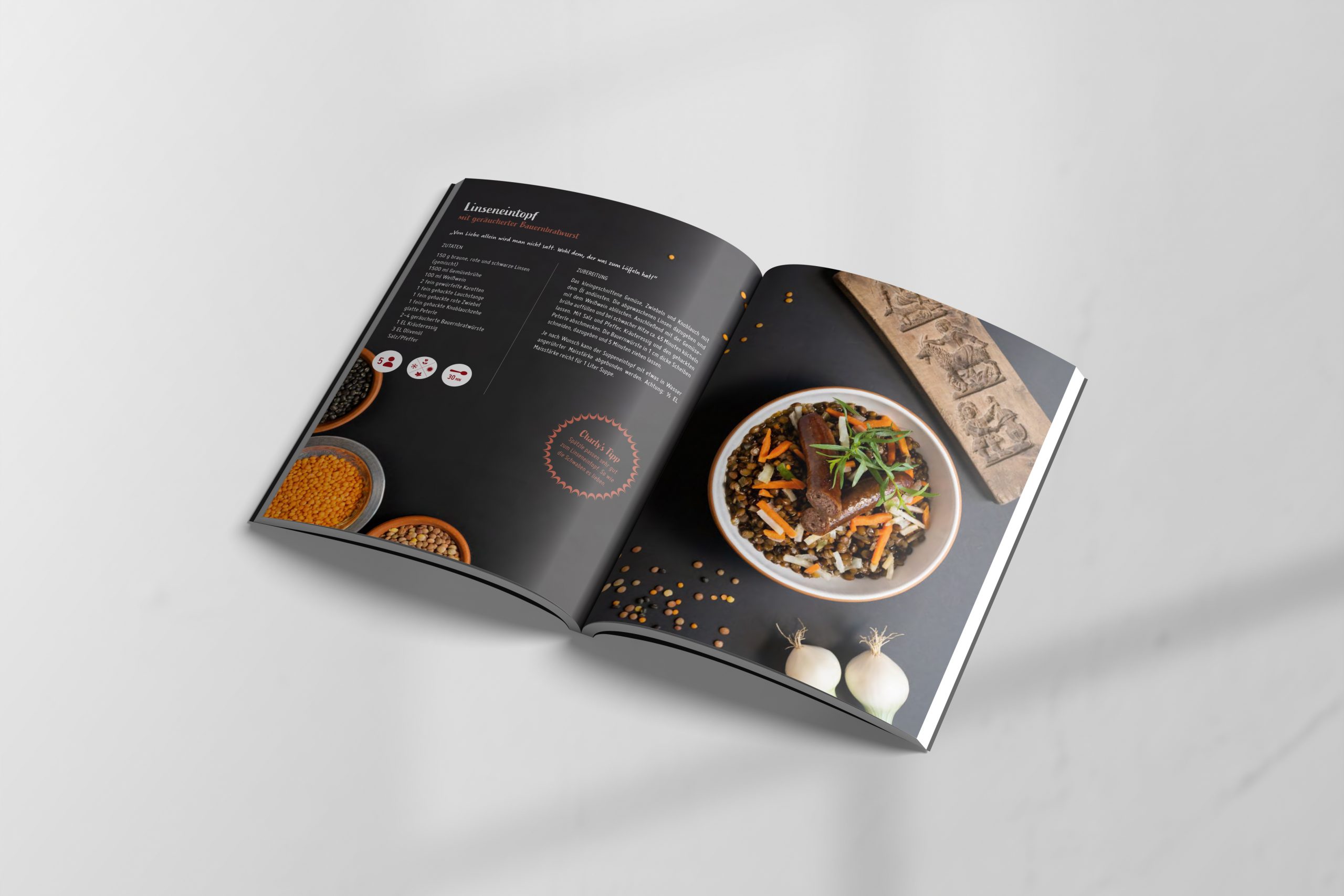 Food-Fotografie: Linseneintopf mit geräucherter Bauernwurst in dunkler Steingutschüssel, arrangiert mit Linsen, Zwiebeln und Karotten auf anthrazitfarbenem Hintergrund; flache Tiefe, warme Lichtakzente, rustikale Holztextur als Bildelement rechts